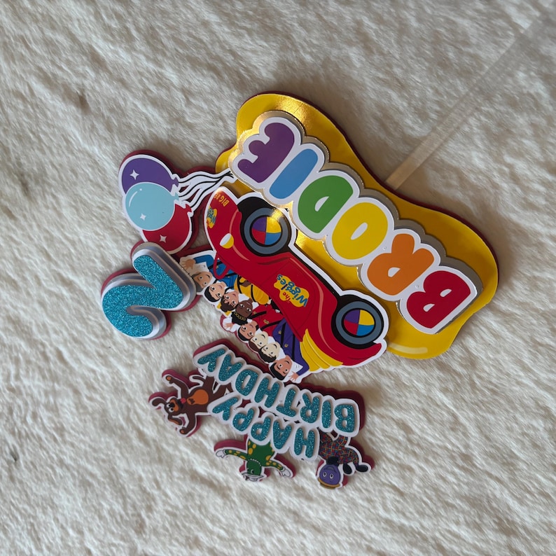 Pu&ograve; includere: Una collezione di decorazioni colorate per torte di compleanno. Include un'auto rossa con la scritta "The Wiggles", il nome "Brodie", il numero "2", palloncini e la frase "Happy Birthday".
