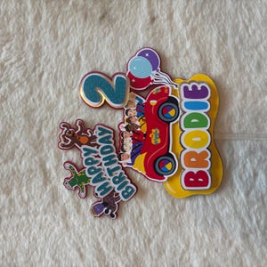 Pu&ograve; includere: Decorazione colorata per torta di compleanno con un'auto rossa, personaggi dei Wiggles, il nome "Brodie" e il numero "2". Include un messaggio "Buon Compleanno" e decorazioni a palloncino. Perfetto per una festa di compleanno per bambini.