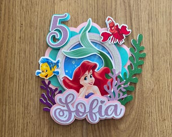 Può includere: Topper per torta a strati con Ariel de La Sirenetta, il numero 5, il nome "Sofia" e immagini dei cartoni animati di Flounder e Sebastian. Il design include accenti rosa, verde acqua e viola.