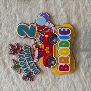 Pu&ograve; includere: Decorazione colorata per torta di compleanno con un'auto rossa con i personaggi dei Wiggles, un numero 2 blu glitterato e il nome "Brodie" in colori arcobaleno. Include il testo "Happy Birthday" e decorazioni con palloncini.