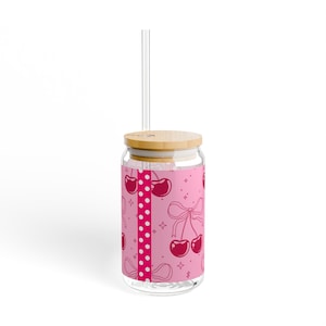 Könnte beinhalten: Ein transparenter Glasbecher mit Bambusdeckel und Glasstrohhalm. Die Außenseite zeigt ein rosa Design mit einem vertikalen weißen Punktstreifen, dunkelrosa Kirschillustrationen, Schleifen und kleinen vierzackigen Sternen als Akzenten.