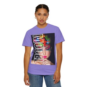 Könnte beinhalten: Ein hellviolettes Kurzarm-T-Shirt mit einem rechteckigen Grafikdruck auf der Brust. Der Druck zeigt ein Gesicht mit lebhaftem, mehrfarbigem Make-up und Blumenschmuck, überlagert mit dem Wort MODEL in großen, fetten weißen Großbuchstaben.