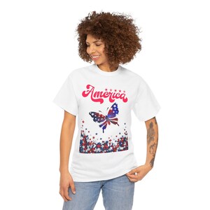 May include: White t-shirt featuring the word "America" in red script above a butterfly design. The butterfly is colored with red, white, and blue, and is surrounded by red and blue stars, creating a patriotic theme.