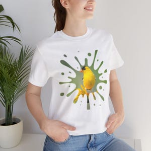 Könnte beinhalten: Ein weißes Kurzarm-T-Shirt mit einem Grafikdruck eines leuchtend gelben Distelfinken mit schwarzer Kappe, der auf einer gelben Blume sitzt, zentriert innerhalb eines großen, unregelmäßigen olivgrünen Farbspritzers auf der Vorderseite.