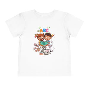 Könnte beinhalten: Ein weißes Kurzarm-T-Shirt mit einer bunten Grafik von zwei Kindern, die gemeinsam ein Buch lesen, umgeben von einem Waschbären, einer Ente und einer Gans, die ebenfalls lesen. Die Buchstaben ABC sind in bunten Farben über den Figuren gedruckt.
