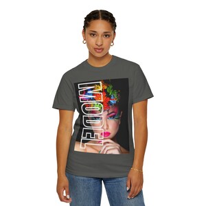 Könnte beinhalten: Ein anthrazitfarbenes Kurzarm-T-Shirt mit einem rechteckigen Grafikdruck auf der Brust. Der Druck zeigt das Porträt einer Person mit lebhaftem, mehrfarbigem Make-up und Haar, überlagert mit dem Wort MODEL in fetten, weißen Großbuchstaben.