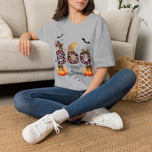 May include: A heather grey short-sleeve t-shirt featuring a graphic print with the text BOO and Happy Halloween. The design includes a leopard print pattern, two orange pumpkins with black bows, a leopard, bats, and a spider web illustration.