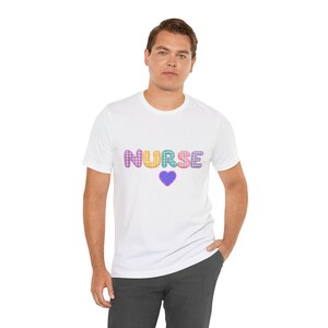 Könnte beinhalten: Ein weißes T-Shirt mit kurzen Ärmeln, auf dessen Brust das Wort NURSE in einem mehrfarbigen Vichy-Karo-Muster gedruckt ist. Unter dem Text befindet sich ein kleines violettes Herz mit weißen Punkten. Das T-Shirt hat einen Rundhalsausschnitt.