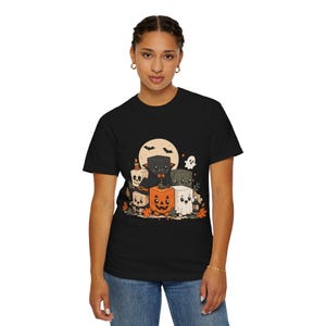 Könnte beinhalten: Ein schwarzes T-Shirt mit einem Grafikdruck verschiedener würfelförmiger Charaktere in Halloween-Kostümen, darunter ein Kürbis, eine Fledermaus, ein Geist und ein Skelett, vor einem blassen Mondhintergrund mit Herbstblättern und kleinen Fledermäusen.