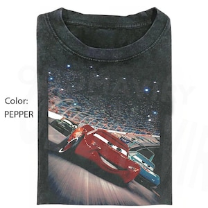 May include: A pepper-colored crew neck t-shirt featuring a graphic print of a red race car and a blue race car driving on a track during a night race with a stadium crowd in the background. The text Color: PEPPER is printed on the left side.