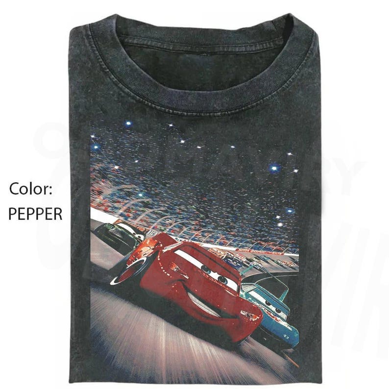 May include: A pepper-colored crew neck t-shirt featuring a graphic print of a red race car and a blue race car driving on a track during a night race with a stadium crowd in the background. The text Color: PEPPER is printed on the left side.