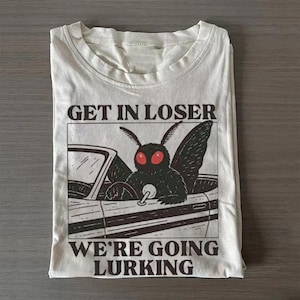 May include: A cream-coloured t-shirt featuring a black graphic of a moth driving a convertible car. The moth has large, bright red eyes. Above the graphic, the text reads GET IN LOSER, and below the graphic, the text reads WE'RE GOING LURKING.