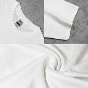 May include: A plain white t-shirt featuring a crew neckline and short sleeves. The garment is made of a solid white fabric with a visible texture. A small, rectangular grey tag is attached to the inside of the collar at the back neckline.