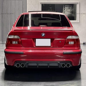 May include: Rear view of a red BMW sedan. The car has a spoiler, dual exhaust pipes, and a black lower bumper. The BMW logo is visible on the trunk. The car is parked indoors.