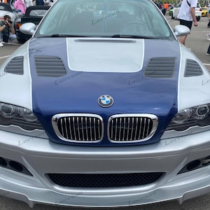 Op de afbeelding: Een zilveren en donkerblauwe BMW-auto met een geventileerde motorkap, een verchroomde niergrille en een voorbumper met geïntegreerde luchtinlaten. Het voertuig toont het BMW-embleem op de motorkap en heeft een tweekleurig lakwerk.