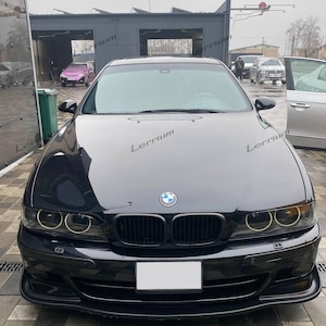 Może przedstawiać: Czarny sedan marki BMW widziany od przodu, wyposażony w charakterystyczny podwójny grill w kształcie nerek, okrągłe reflektory typu halo oraz centralnie umieszczony niebiesko-biały emblemat marki na masce. Pojazd ma lśniące wykończenie i pustą tablicę.