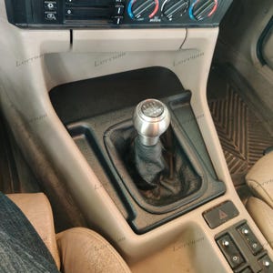 Könnte beinhalten: Ein silberner Schalthebelknauf mit schwarzer Lederhülle, eingebaut in der Mittelkonsole eines Autos. Der Knauf zeigt ein Diagramm des Gangmusters. Die Konsole ist beige und schwarz, mit verschiedenen Knöpfen und Bedienelementen.