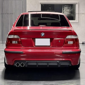 May include: Rear view of a red BMW sedan. The car has a spoiler, dual exhaust pipes, and black trim. The BMW logo is visible on the trunk. The car is parked indoors.