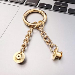Pode incluir: Porta-chaves de metal dourado com um anel grande e duas correntes. Cada corrente termina com uma peça pequena e redonda de cor dourada. O porta-chaves está sobre uma superfície cinzenta.