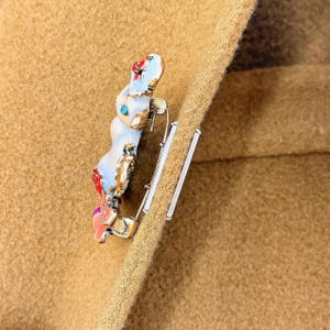 May include: A colorful enamel bunny brooch with a silver-tone pin. The bunny is white with blue, red, and pink accents. The brooch is attached to a tan fabric, showcasing the jewelry's design and color contrast.