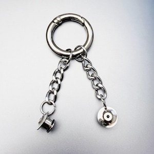 Pode incluir: Acessório metálico prateado com um grande fecho circular, duas correntes, uma pequena rolha cilíndrica e um pingente plano e circular. As correntes pendem do fecho. O item tem um acabamento metálico brilhante.