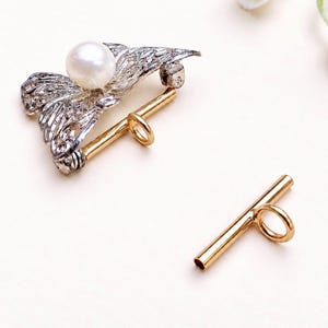 May include: A toggle clasp set featuring a silver-toned filigree component adorned with a single white pearl and a matching gold-toned metal bar. The intricate silver piece includes a loop for attachment, while the gold bar has a central ring.