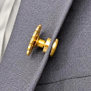 May include: A gold-toned metal lapel pin featuring a textured circular front and a smooth cylindrical post, secured through the fabric of a dark grey blazer. The pin has a polished finish and is attached to the lapel with a matching gold-toned backing.