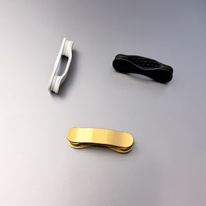 以下が含まれることがあります： 白、黒、金の3つのマグネットクリップ。各クリップは、滑らかで細長い曲線的なデザインで、両端が丸みを帯びています。白いクリップは垂直に配置され、黒と金のクリップは水平に向けられています。