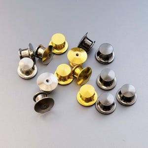 May include: A collection of pin backs in various metallic finishes, including gold, silver, and gunmetal. The pin backs are round with a flat base and a post for attaching to a pin. The image is taken on a gray surface.