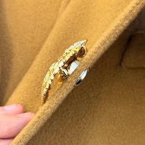 Może przedstawiać: Złota broszka w kształcie ptaka, ozdobiona małymi, przezroczystymi kamieniami. Broszka jest przymocowana do beżowej tkaniny, z widocznym w pobliżu srebrnym pierścieniem. Ptasi motyw ma szczegółowe wzory skrzydeł i piór.