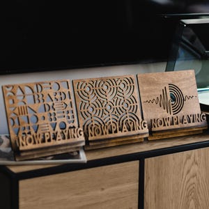 May include: Three brown wooden display stands for vinyl records, each featuring the text "NOW PLAYING" at the base. The stands have intricate laser-cut designs, including geometric patterns, concentric circles, and a stylized sound wave graphic.