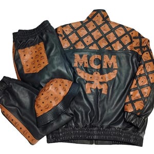 May include: A black and cognac brown leather jacket, matching pants, and a bucket hat set. The jacket features the MCM logo, a large MCM text appliqu&eacute; on the back, and a diamond grid pattern. The pants have a cognac pocket and side stripes.