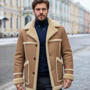 May include: A tan shearling coat with a cream-colored shearling collar and trim. The coat has a button-up front, two front pockets, and a classic design. The person in the image is wearing a navy blue turtleneck and gray pants.