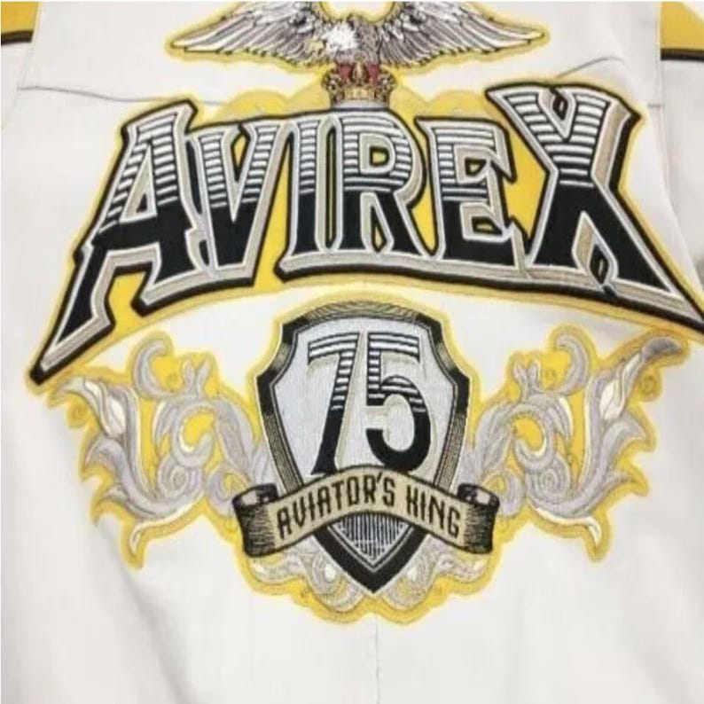 May include: Close-up of a white jacket with embroidered details. The jacket features the brand name "AVIREX" in a bold, stylized font, with a yellow outline and black fill. Below, a shield displays the number "75" and the text "AVIATOR'S KING". An eagle with a crown is at the top.