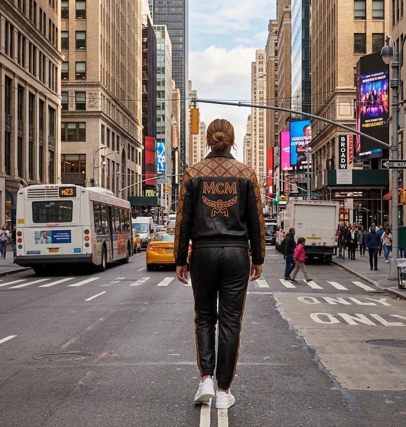 May include: A black leather jacket with brown patterned sleeves and the MCM logo in orange on the back, paired with matching black leather joggers featuring a side stripe and white sneakers, worn in a busy city street setting.