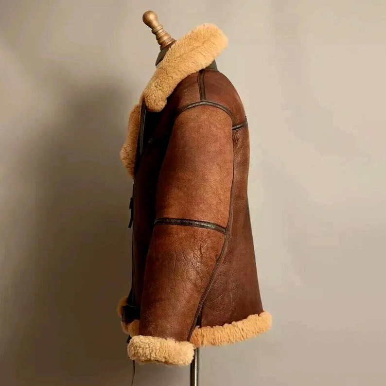 May include: A brown leather jacket with a tan shearling collar and trim. The jacket is displayed on a mannequin, showcasing its side profile. The jacket has a classic design with visible stitching and a warm, inviting aesthetic.