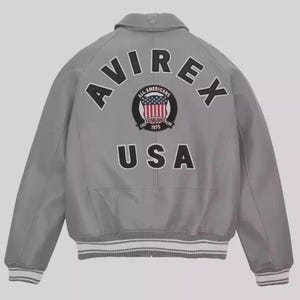 May include: A grey bomber jacket featuring the word "AVIREX" in black lettering across the back. Beneath this is a circular emblem with "ALL AMERICANS ESTABLISHED 1975" and a USA flag design. The word "USA" is printed below the emblem.