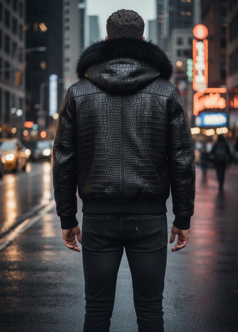 May include: A black leather jacket with a textured, crocodile-like pattern and a fur-lined hood. The jacket has a bomber-style design with a ribbed hem and cuffs. The person is wearing black jeans. The background shows a city street with blurred lights.