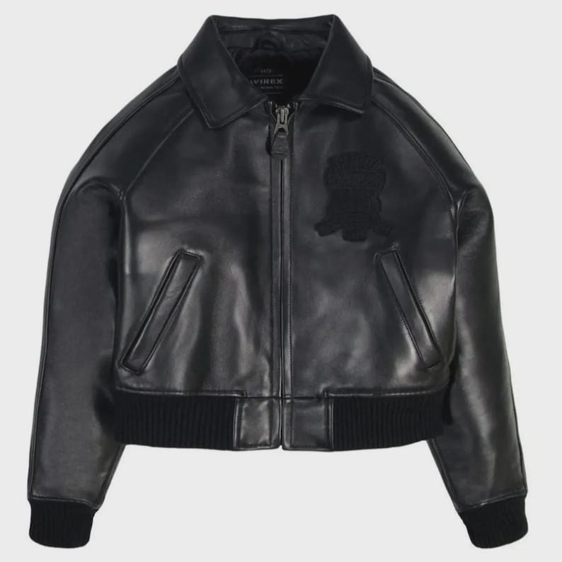 May include: Black leather jacket with a zip-up front, collar, and ribbed cuffs and hem. Features a patch on the chest. The jacket has two front pockets and the brand name "AVIREX" is visible on the collar.
