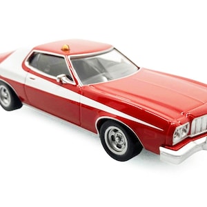 May include: A red and white die-cast model car with a white stripe along the side. The model car has a red body, a white roof, and chrome accents. The car has black tires and a small amber light on the roof.