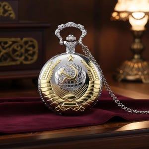 May include: Silver and gold-toned pocket watch with a chain. The watch face features a hammer and sickle emblem over a globe, surrounded by wheat and a rising sun design. The watch has a decorative handle and a silver chain.