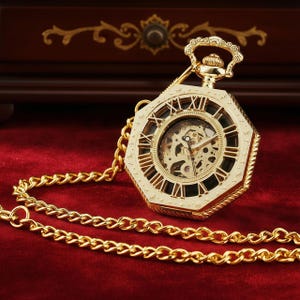 May include: A gold-colored, octagonal pocket watch with Roman numerals and a visible inner mechanism. The watch is attached to a gold chain, resting on a red velvet surface. The watch has a decorative crown and a detailed design.