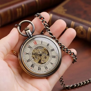 Op de afbeelding: Antieke zakhorloge met een bronzen behuizing en ketting. De wijzerplaat toont Romeinse cijfers en een zichtbaar mechanisch uurwerk. Het horloge wordt in een hand gehouden, wat het ingewikkelde ontwerp benadrukt.