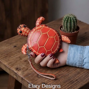 Könnte beinhalten: Eine handgefertigte, orangefarbene Schildkröten-Geldbörse mit Reißverschluss und Handschlaufe. Die Geldbörse hat ein geometrisches Muster auf ihrem Panzer. Der Text "Elary Designs" ist sichtbar.