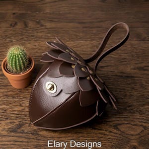 May include: A brown leather coin purse with a unique design, featuring petal-like layers and a handle. The purse has a gold-colored clasp and is next to a small potted cactus. The text "Elary Designs" is at the bottom.