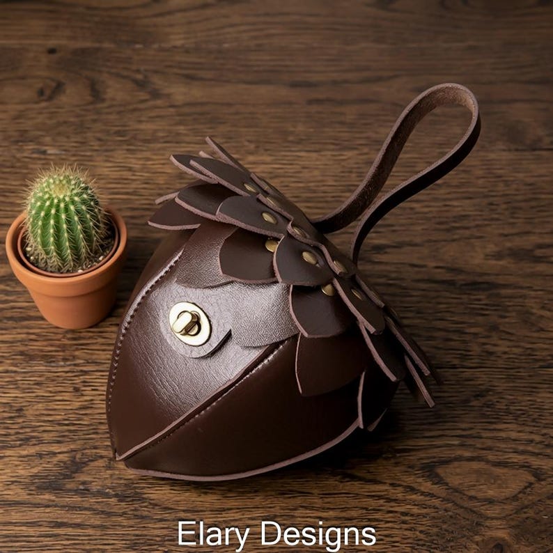 May include: A brown leather coin purse with a unique design, featuring petal-like layers and a handle. The purse has a gold-colored clasp and is next to a small potted cactus. The text "Elary Designs" is at the bottom.