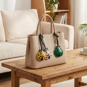 May include: A beige leather tote bag with a structured design. The bag features two handles and is adorned with three ladybug charms in yellow, pink, and green. The ladybugs have black spots and are attached with black straps.