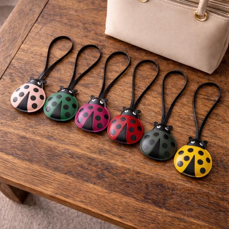 May include: A collection of six ladybug-shaped keychains in various colors, including pink, green, purple, red, dark green, and yellow. Each keychain has a black leather strap and a black body with contrasting spots. A beige handbag is in the background.