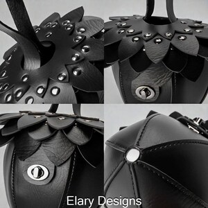 May include: A black leather handbag with a unique design. The bag features petal-like accents around the top and is adorned with silver-toned rivets and a clasp closure. The text "Elary Designs" is visible at the bottom.
