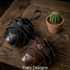 May include: Two leather keychains, one black and one brown, with a unique layered design resembling a pinecone. Each keychain has a small loop handle. A small potted cactus sits nearby. The text "Elary Designs" is at the bottom.